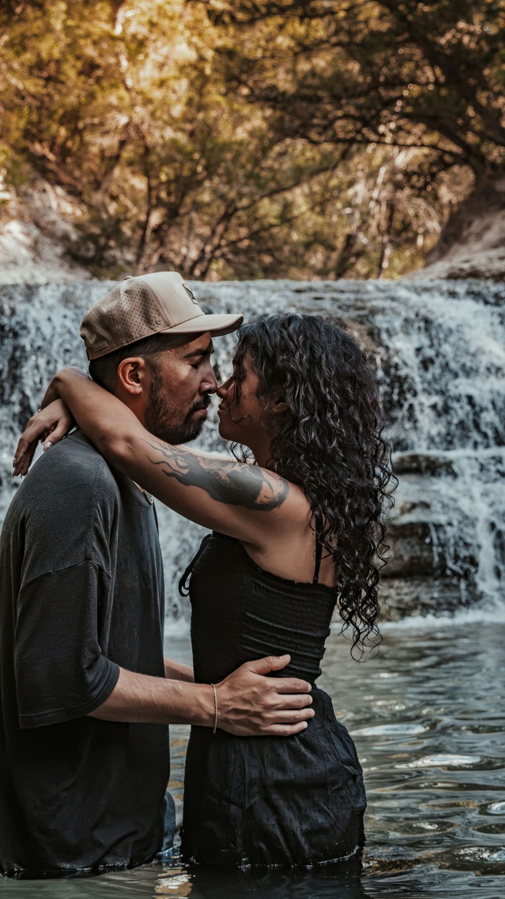 Couple embracing and about to kiss while standing in water before a scenic waterfall outdoors.