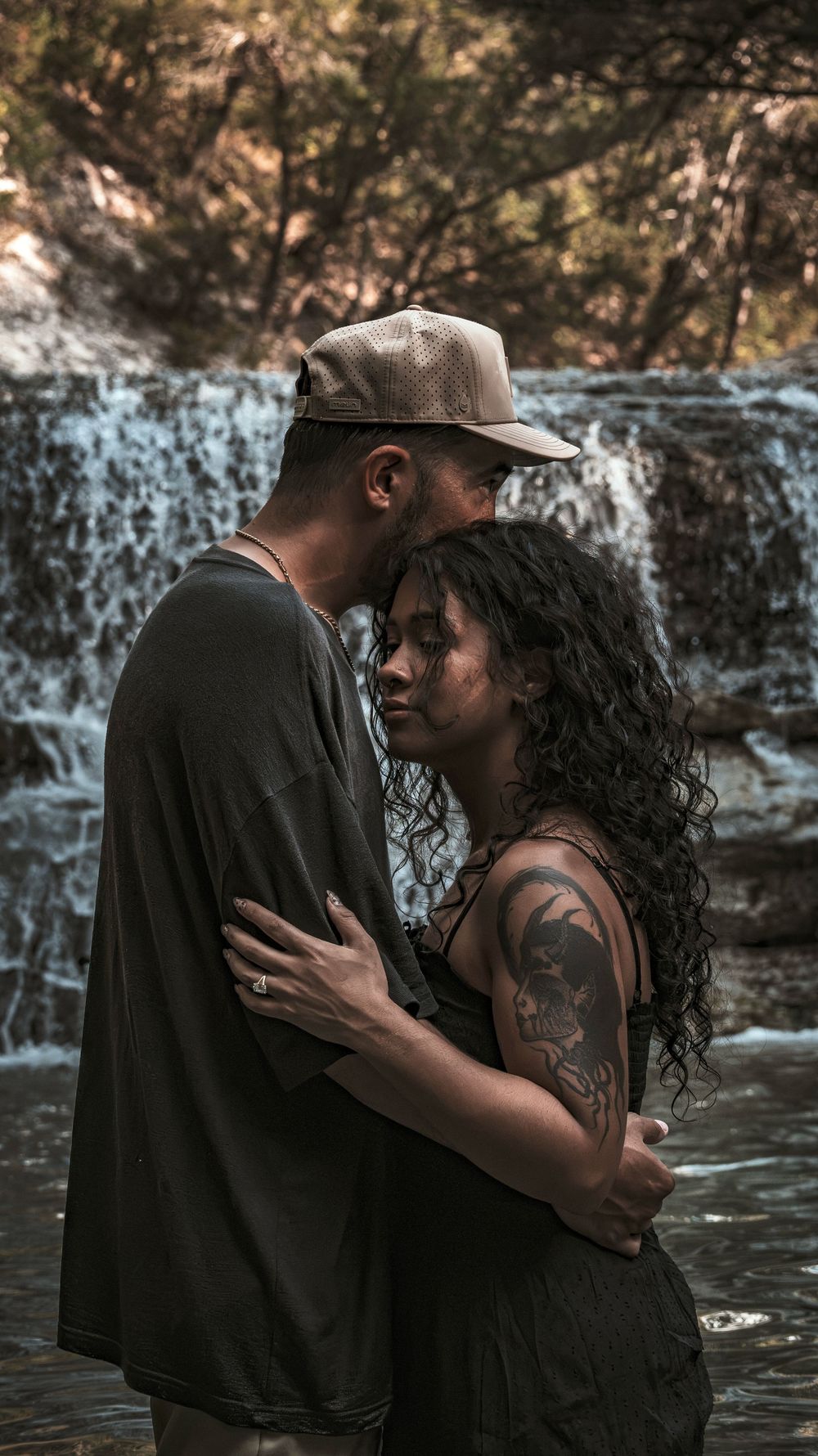 A couple hugging tenderly in shallow water with a scenic waterfall backdrop in a forest setting.