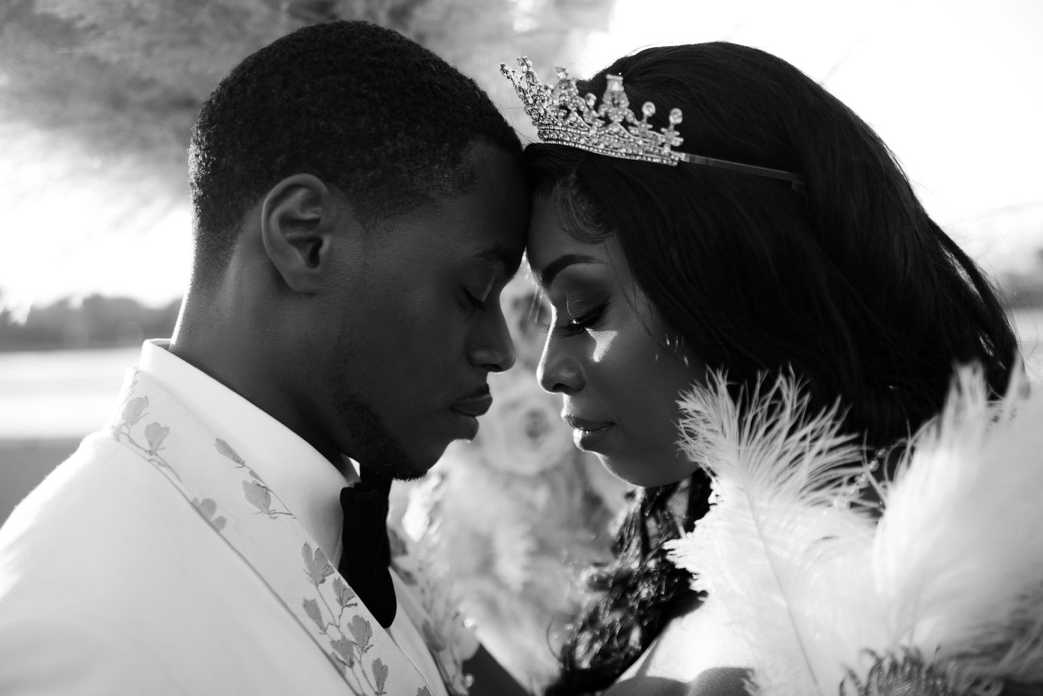 A romantic black and white wedding portrait featuring a couple in elegant attire with a tiara and feathers.