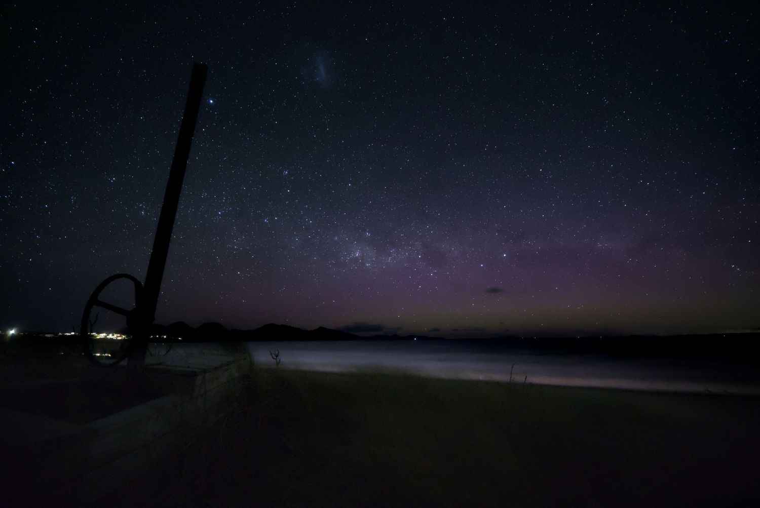 Stunning night sky with Milky Way and purple aurora over a dark beach with a sculpture silhouette.