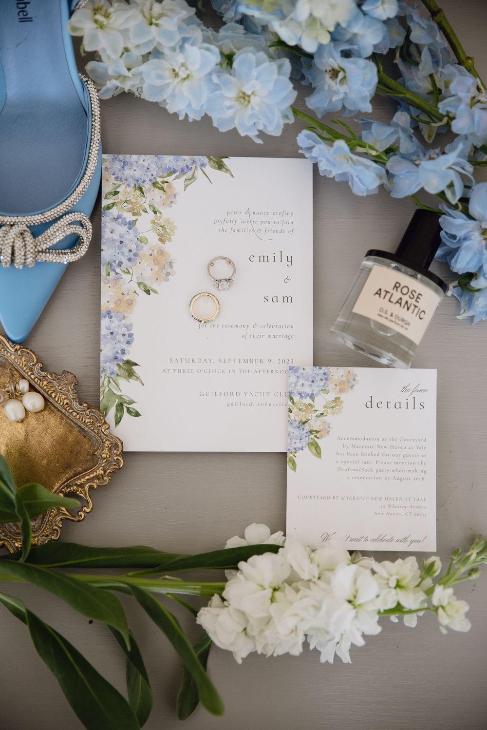Wedding invitation with floral design surrounded by blue shoes, flowers and perfume bottle.