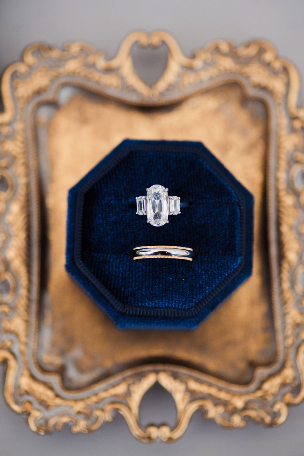 Elegant engagement ring and wedding band displayed in navy blue octagonal box on ornate gold frame.