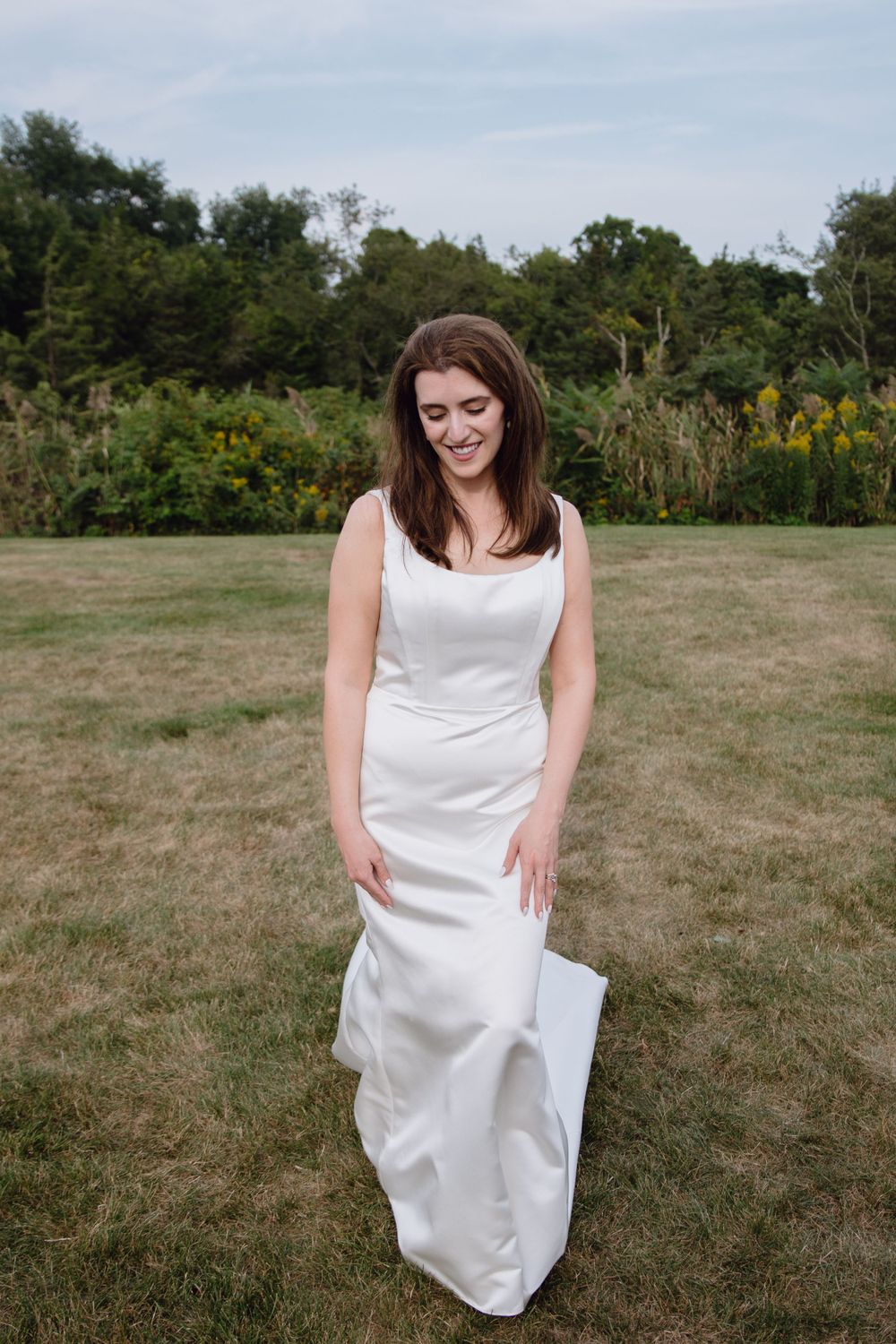 A radiant woman in a white wedding dress stands on a grassy lawn with wildflowers in the background.