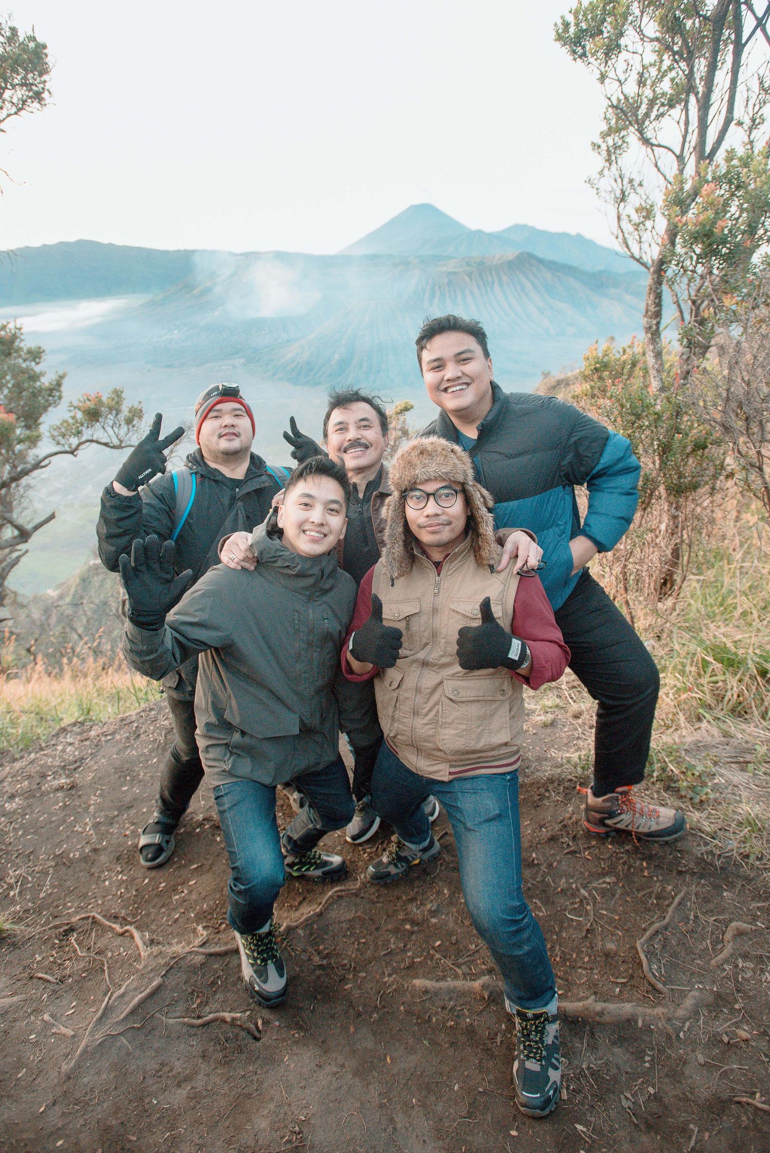 Group of five friends posing happily at Mount Bromo volcano viewpoint, wearing winter jackets in misty mountain landscape.