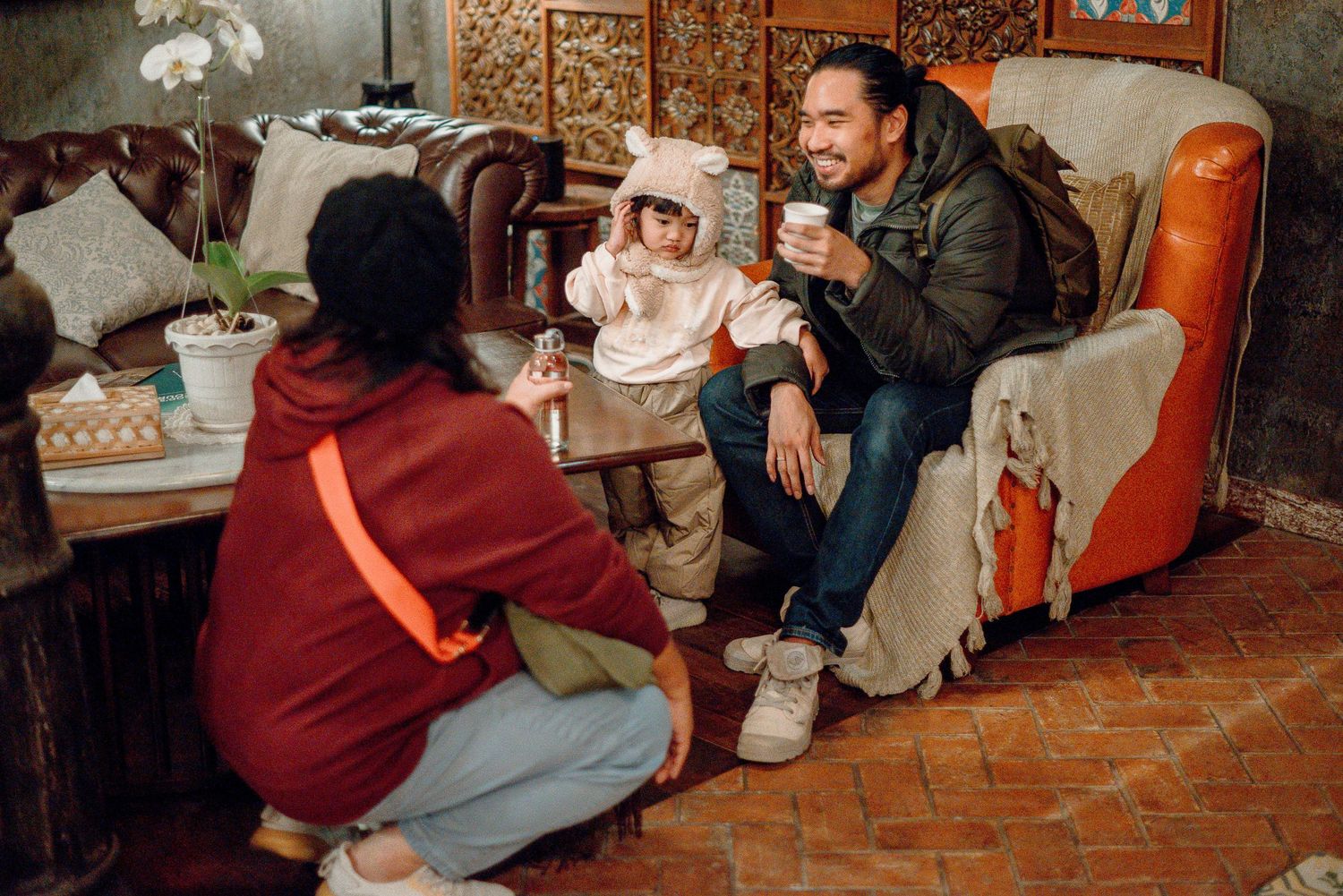 A family with a toddler in a bear hat enjoys drinks together on a cozy vintage couch indoors.