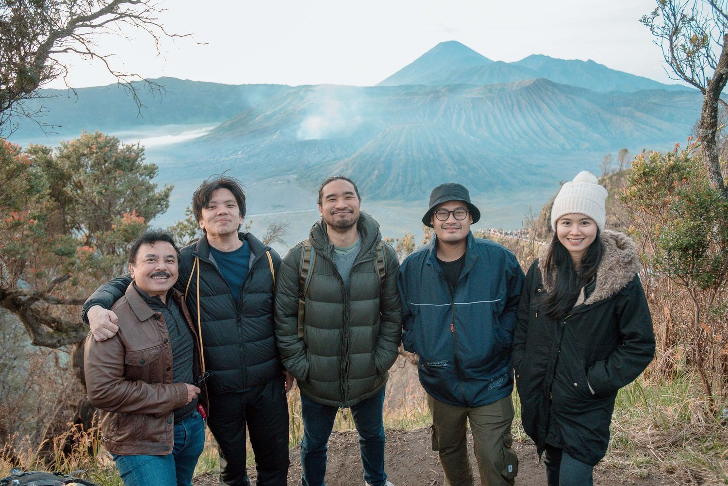 Five friends pose smiling before Mount Bromo volcano crater landscape in East Java Indonesia on a cool misty day.