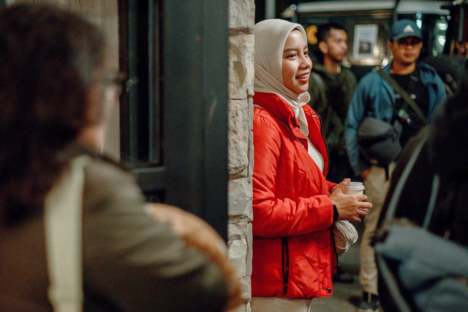 Smiling woman in red jacket and hijab holding coffee cup outdoors in a busy urban setting.