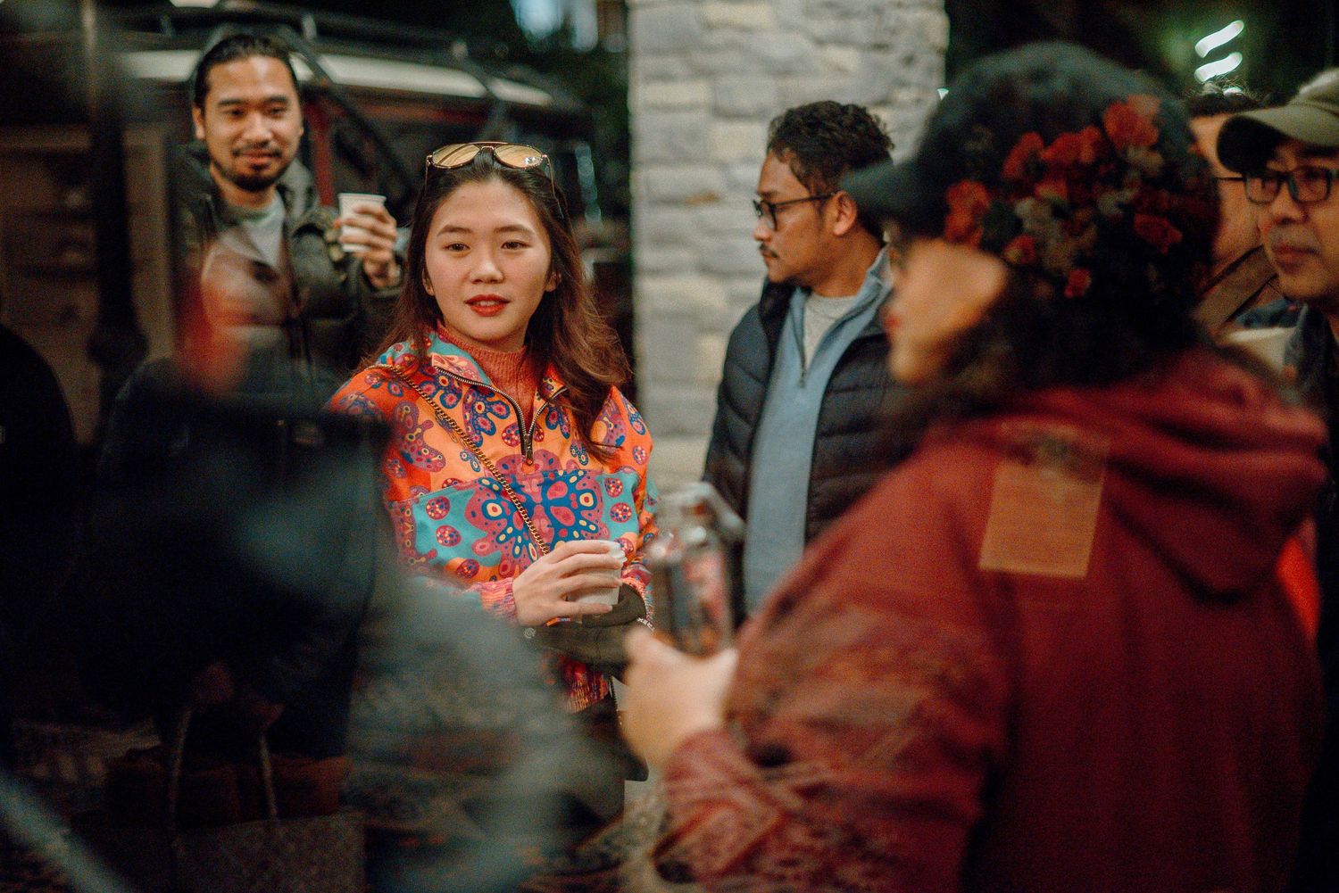 A smiling woman in a colorful floral jacket holds a drink at a festive evening social gathering.