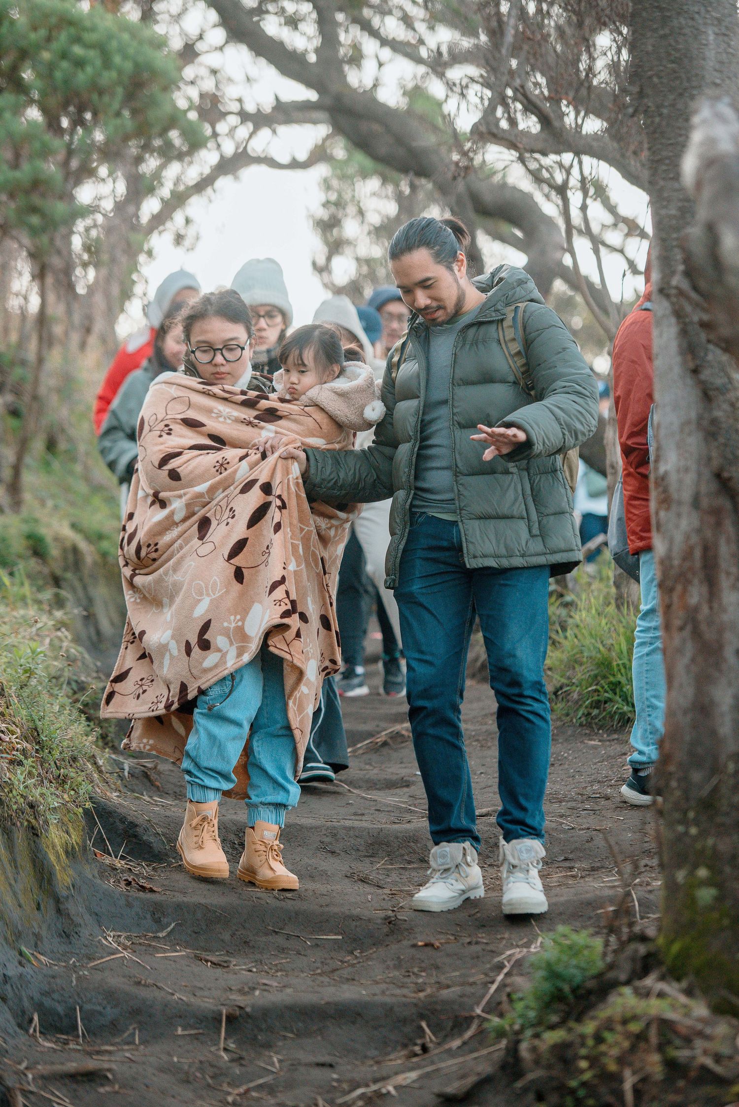 A man in a green jacket shows his phone to a woman wrapped in a blanket on a misty forest trail.