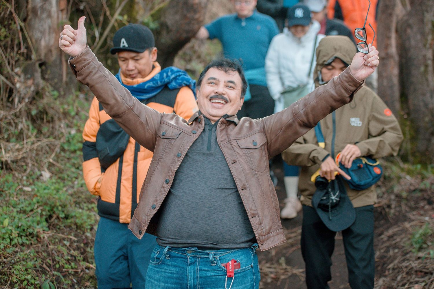 Cheerful man raises both arms in victory on a forest hiking trail with friends in outdoor gear behind him.