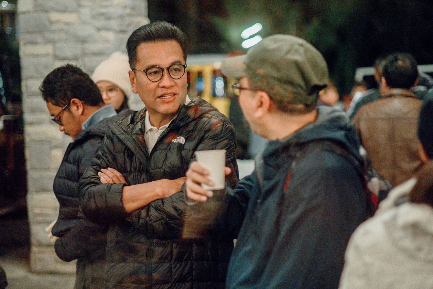 Group of people socializing outdoors at night, man in North Face jacket with arms crossed talking to others.