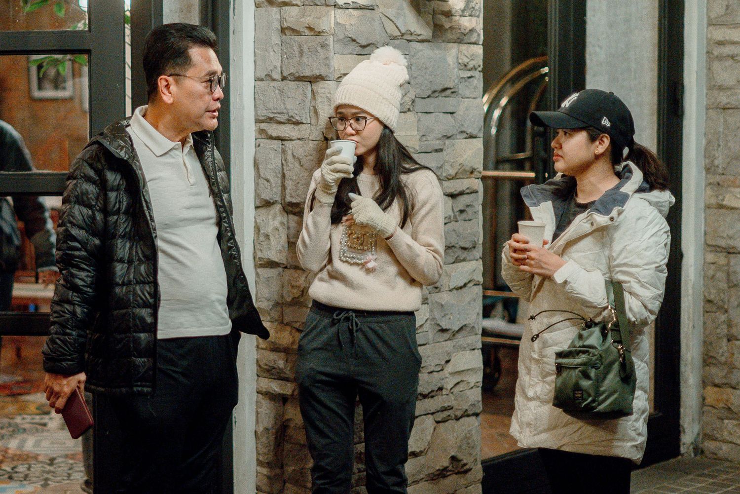Three people in winter coats chat outdoors, two women holding coffee cups, near a stone building at night.