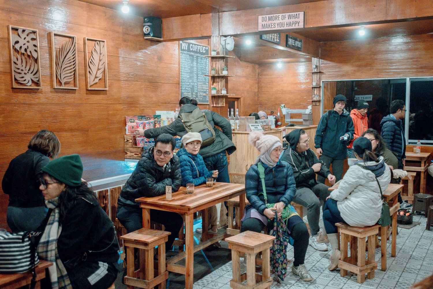 Cozy restaurant interior with wooden furniture, patrons dining in warm lighting, wall art, and a bookshelf in the background.