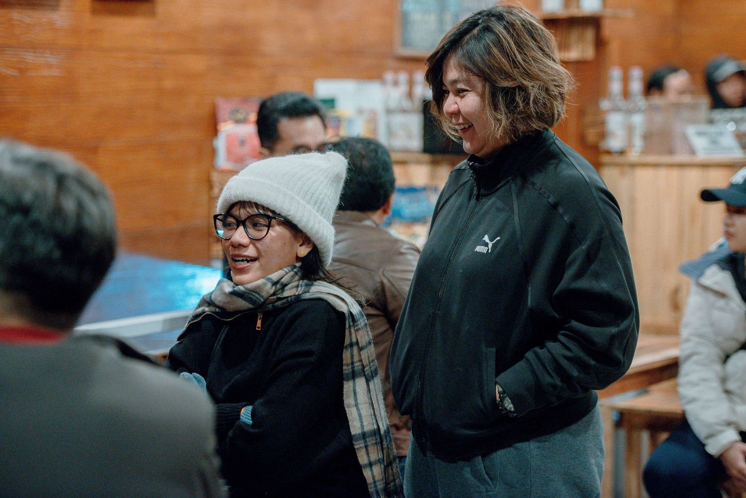 Two women smiling and chatting indoors, one wearing a gray beanie and scarf, the other in a black Puma jacket.