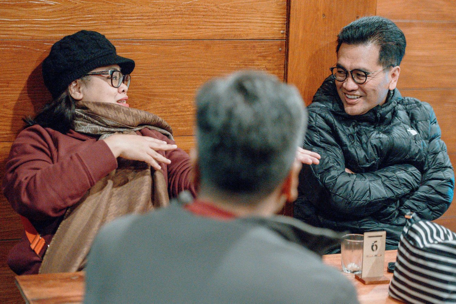 Three friends having a lively conversation in a cozy restaurant booth, wearing winter jackets and glasses.