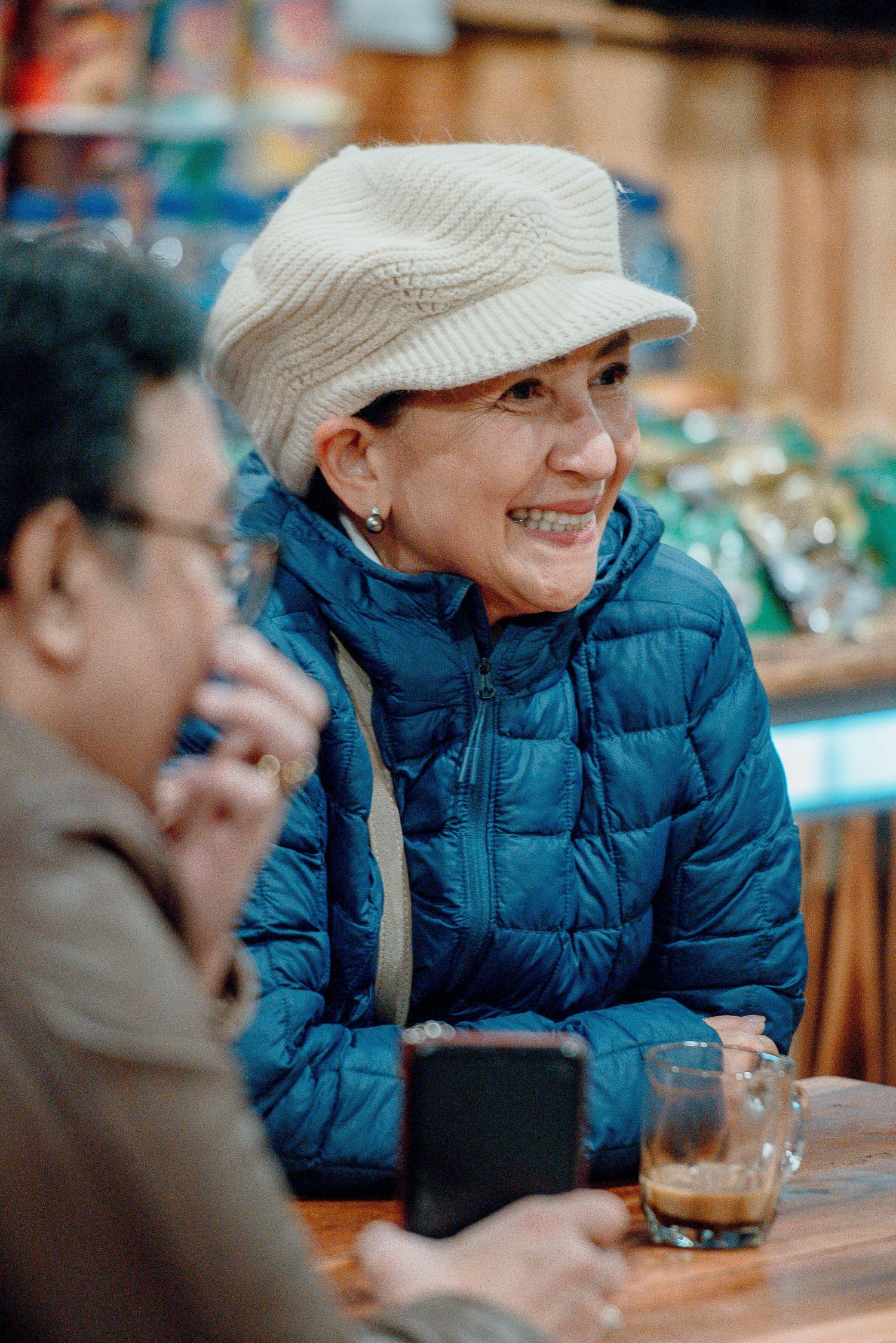 Smiling senior woman in blue puffer jacket and white knit cap enjoying coffee at a cafe table with a companion.