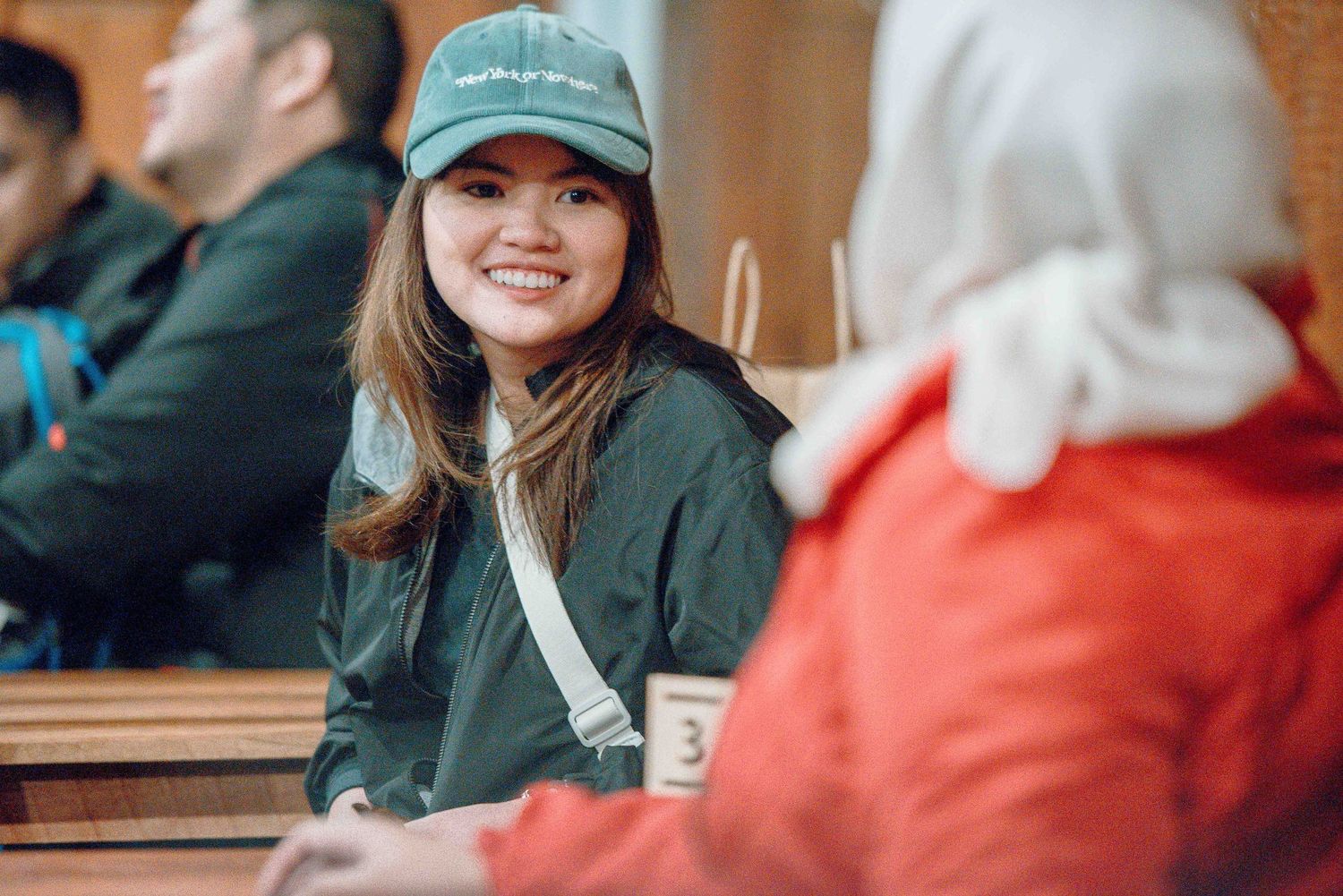 Smiling young woman in green cap and jacket sitting at a table, engaged in conversation indoors.