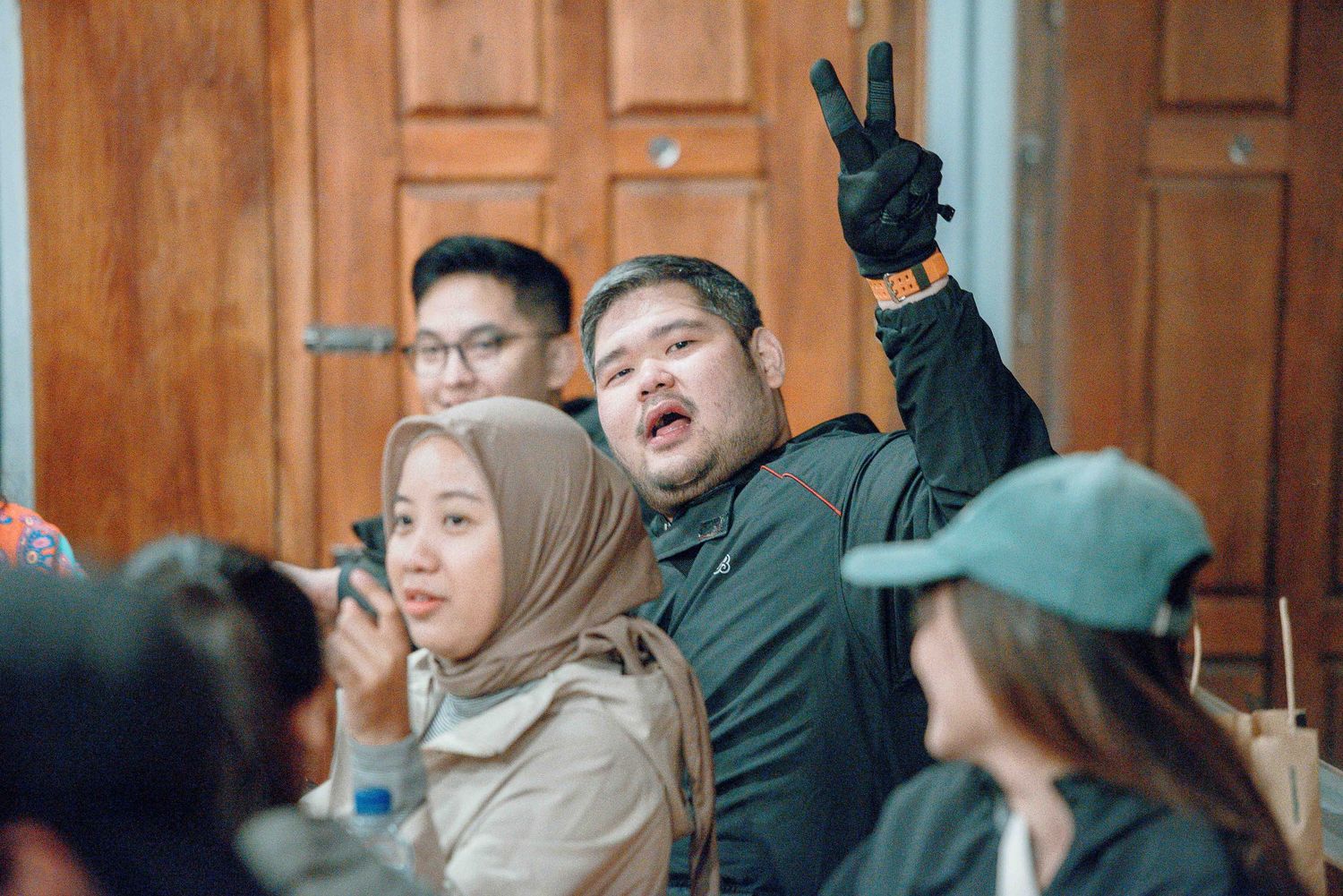 A man flashing a peace sign with people around him indoors near wooden doors.