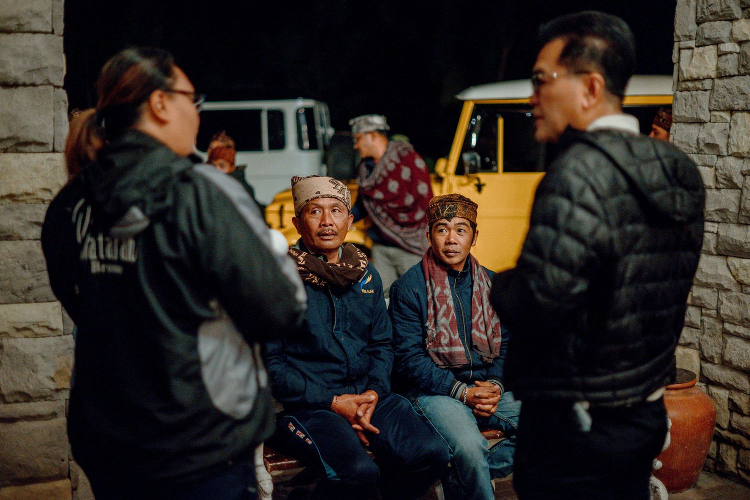 Two men wearing traditional patterned hats sit outdoors at night near vintage yellow vehicles.
