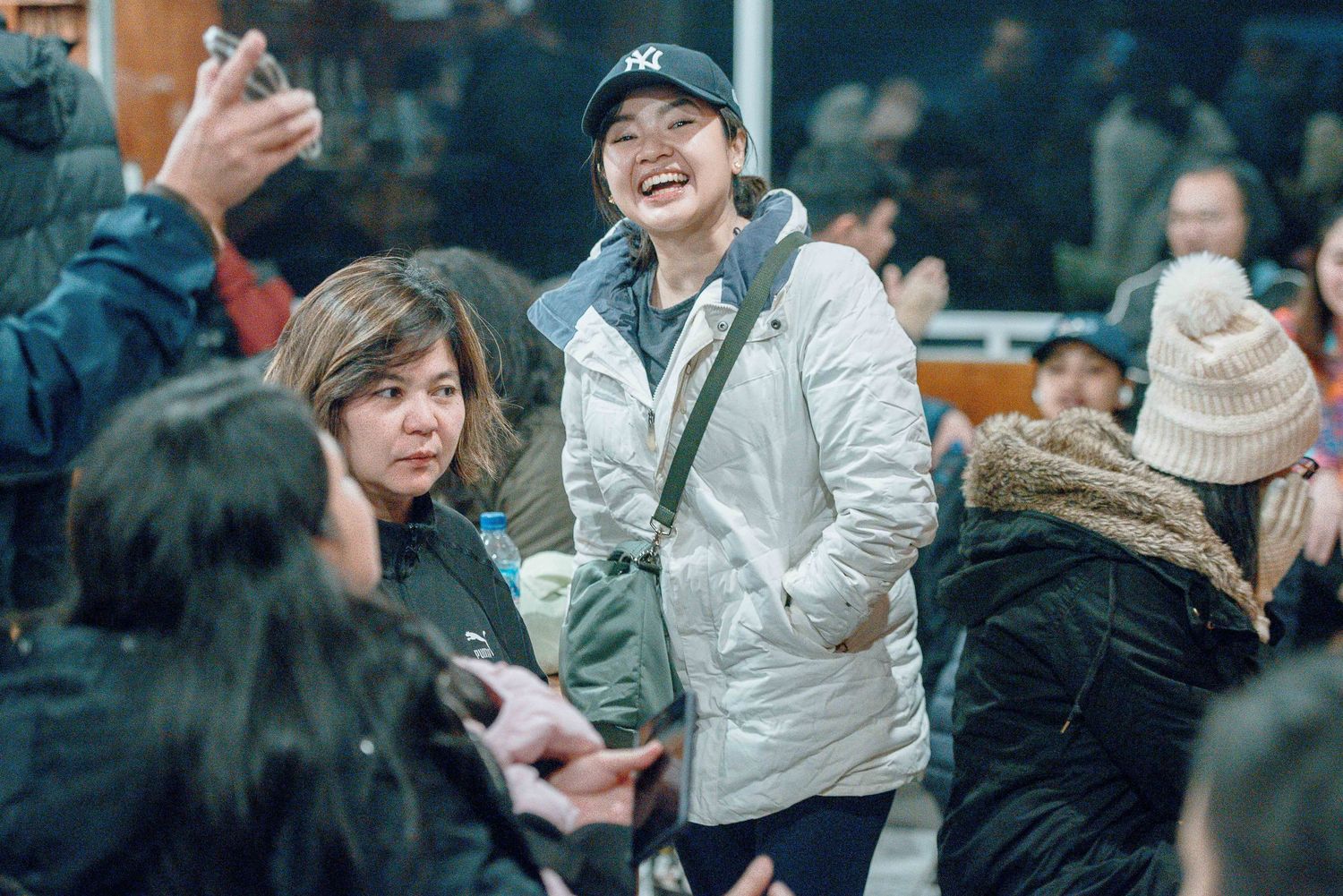 Smiling person in white jacket and NY cap mingles with crowd at an outdoor winter gathering.