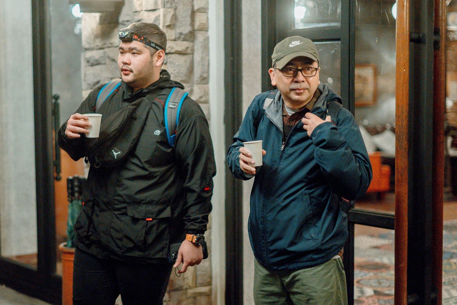 Two men in outdoor jackets holding coffee cups walk together near a building entrance at night.