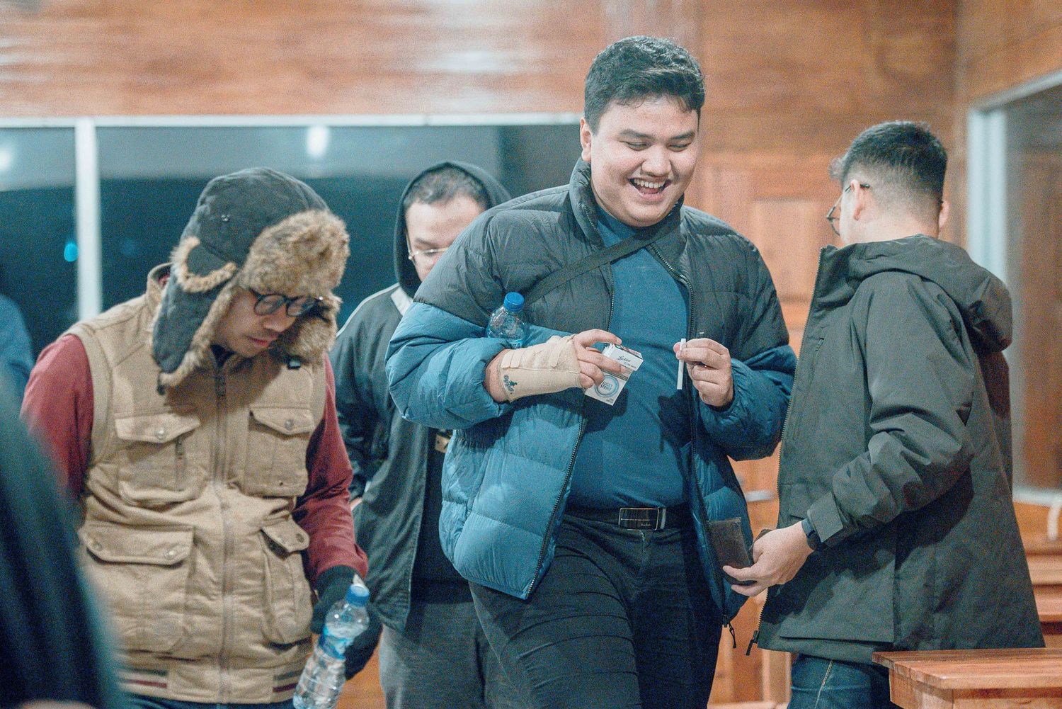 Group of young men smiling and socializing indoors, one wearing a blue puffer jacket holding a small item.