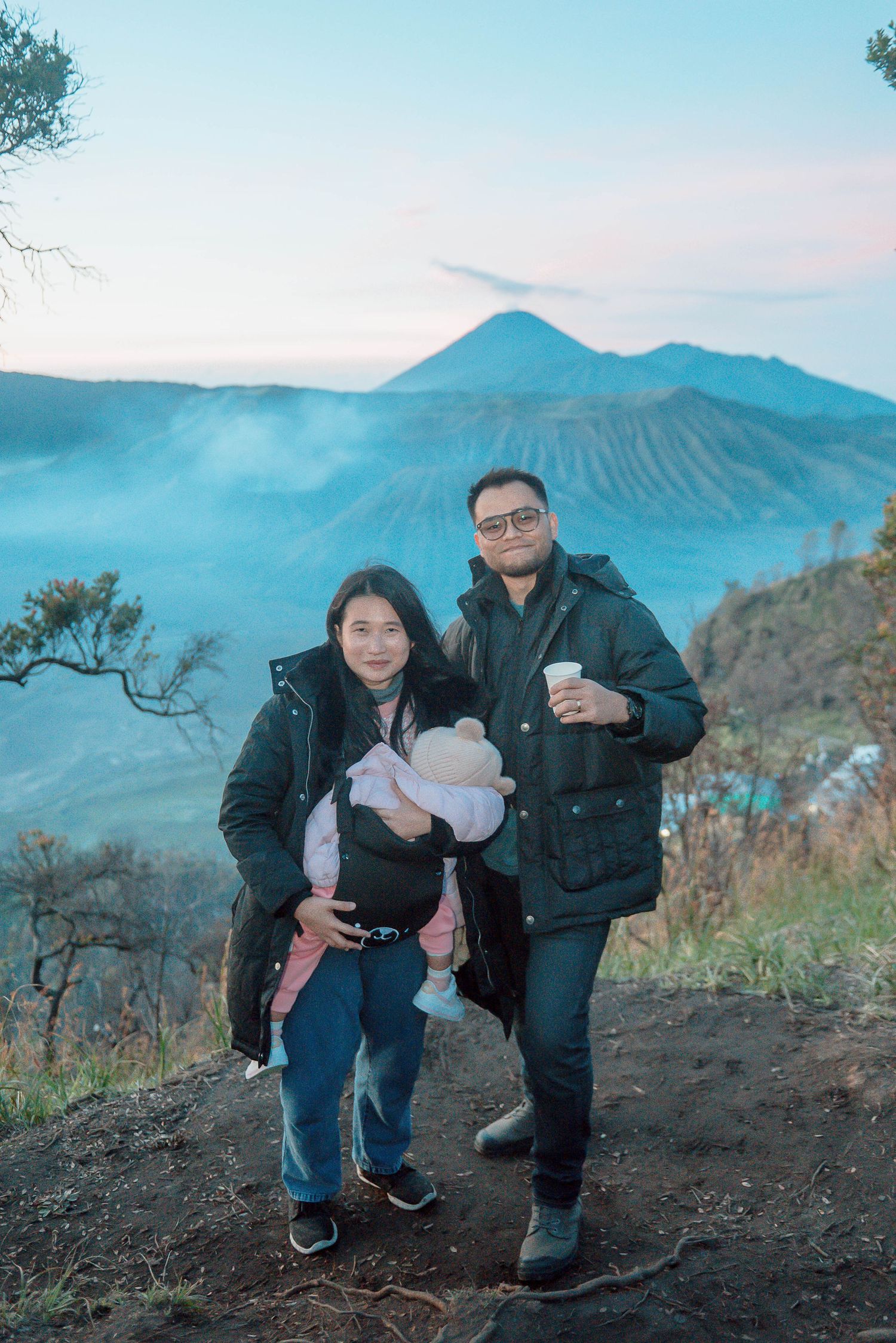 Family posing at Mount Bromo volcano viewpoint in Indonesia at sunrise, with misty volcanic landscape behind.