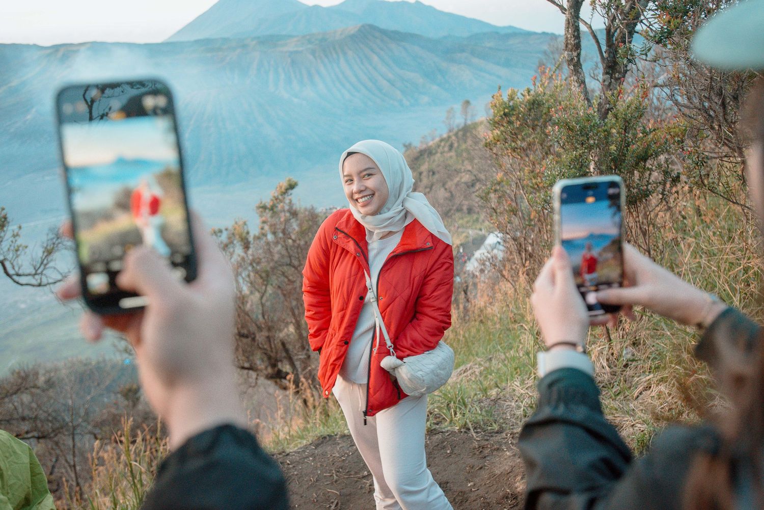 Woman in red jacket and white hijab poses for photos on a mountain trail with volcanic landscape backdrop.