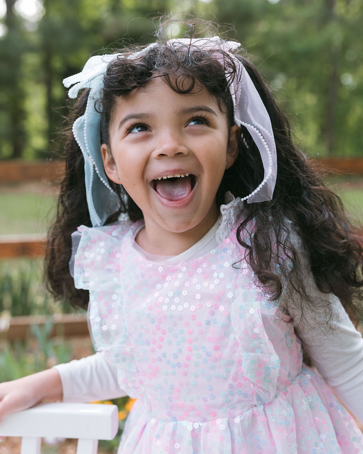 A joyful young girl in a pink sequin dress and white bow laughs outdoors in a lush green garden setting.