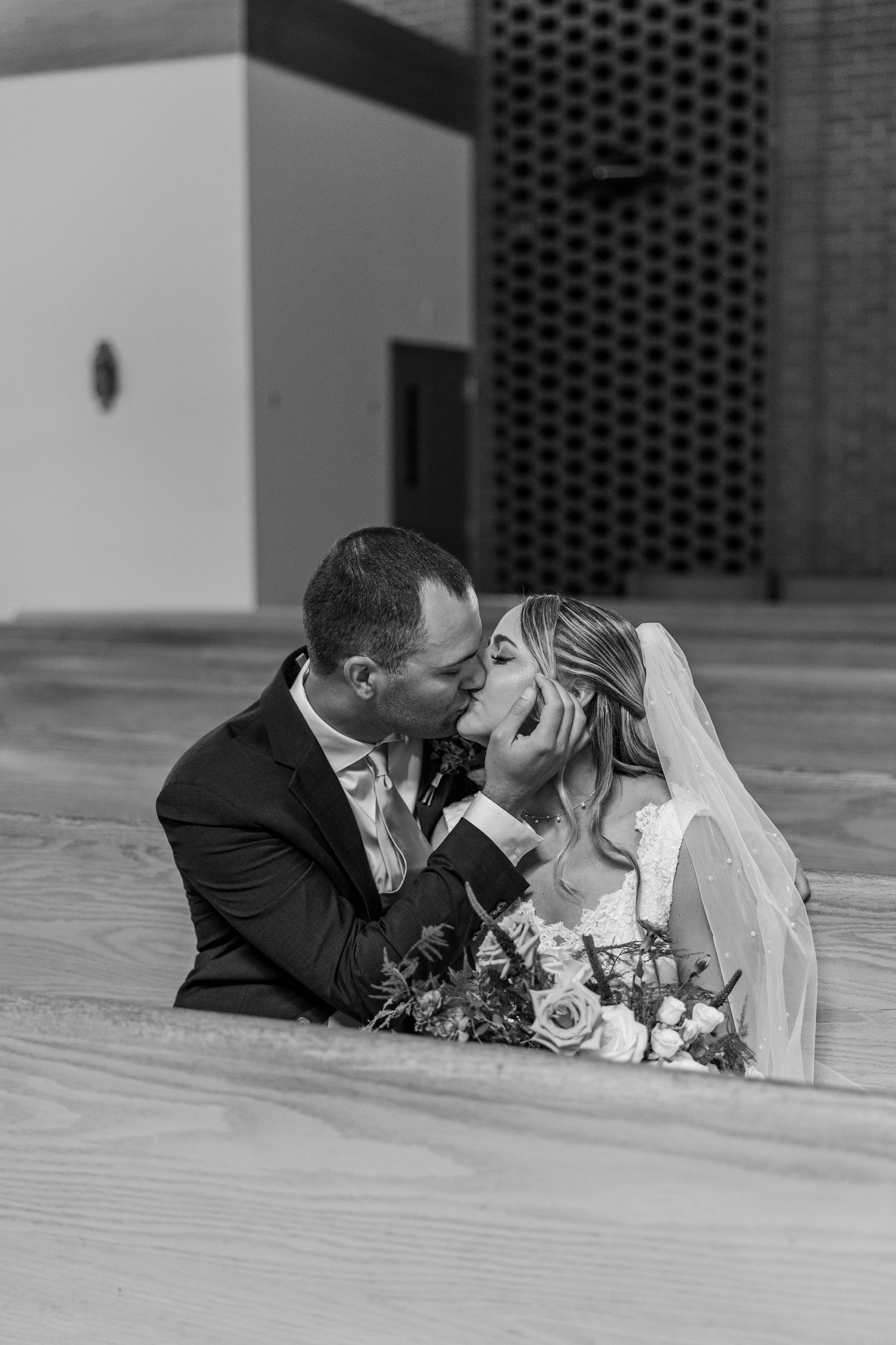 Black and white photo of a bride and groom sharing a kiss while sitting on steps, bride holding a bouquet.