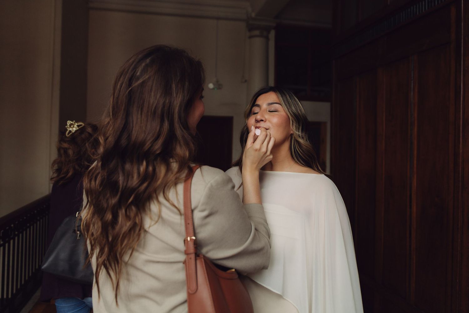 Woman applying makeup to smiling bride in elegant white dress in dimly lit indoor setting.