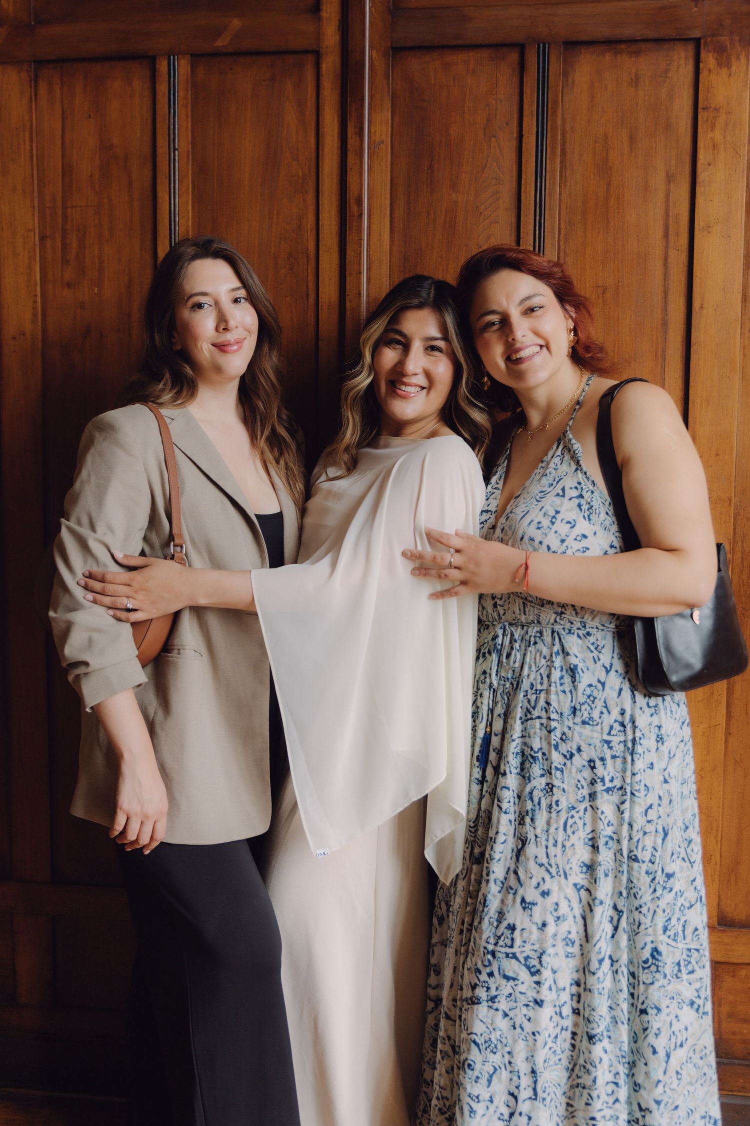 Three women posing together smiling in front of ornate wooden wall panels indoors.