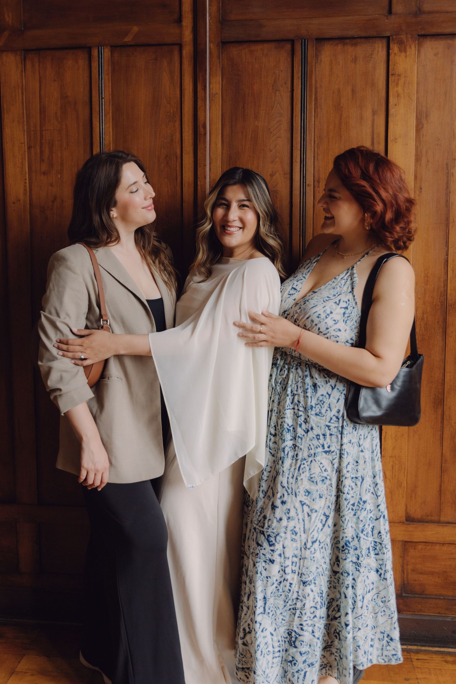 Three women laughing together against wooden paneled wall, bride in white dress with friends.