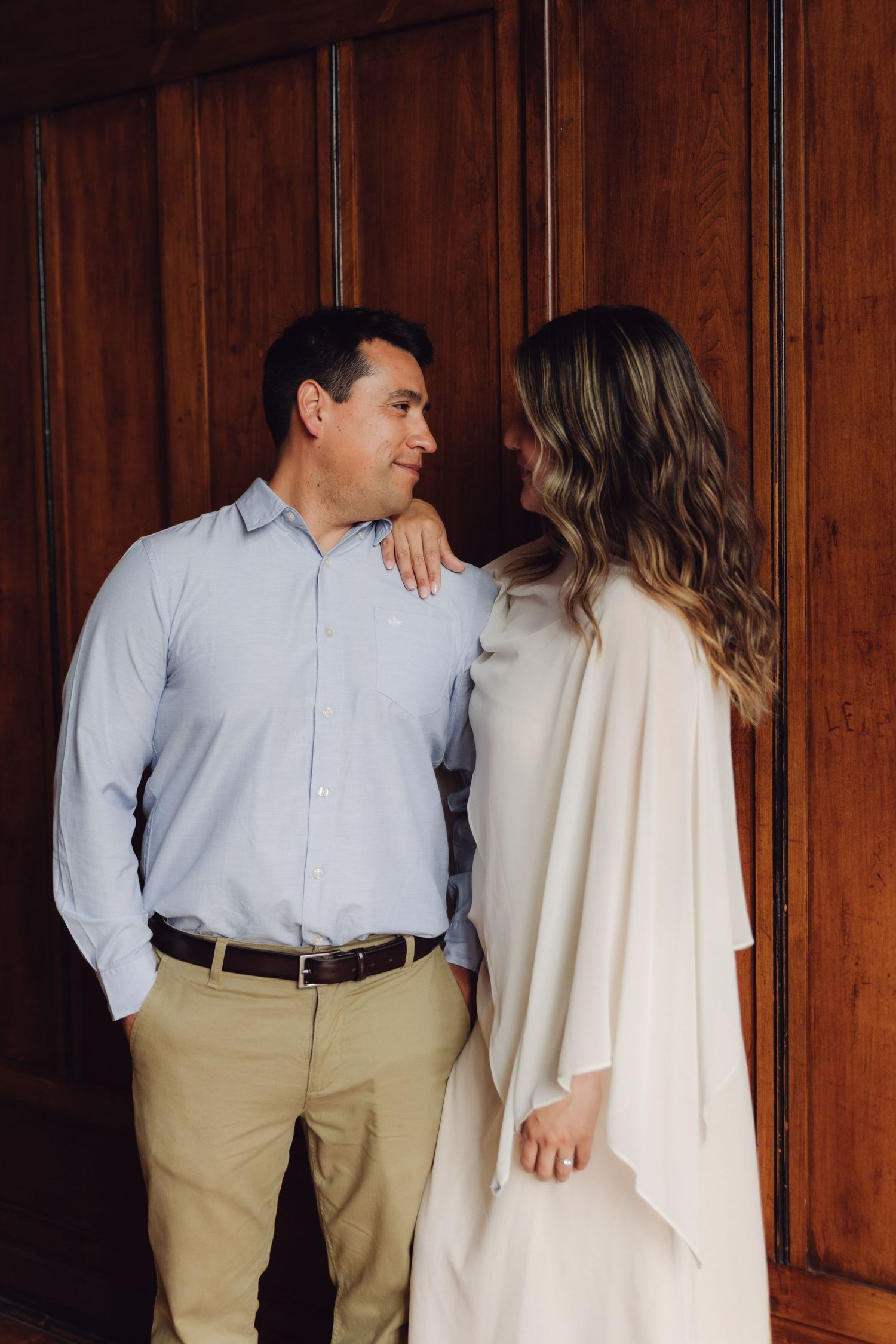 Couple sharing tender glance while standing against rich wooden wall in elegant indoor setting.
