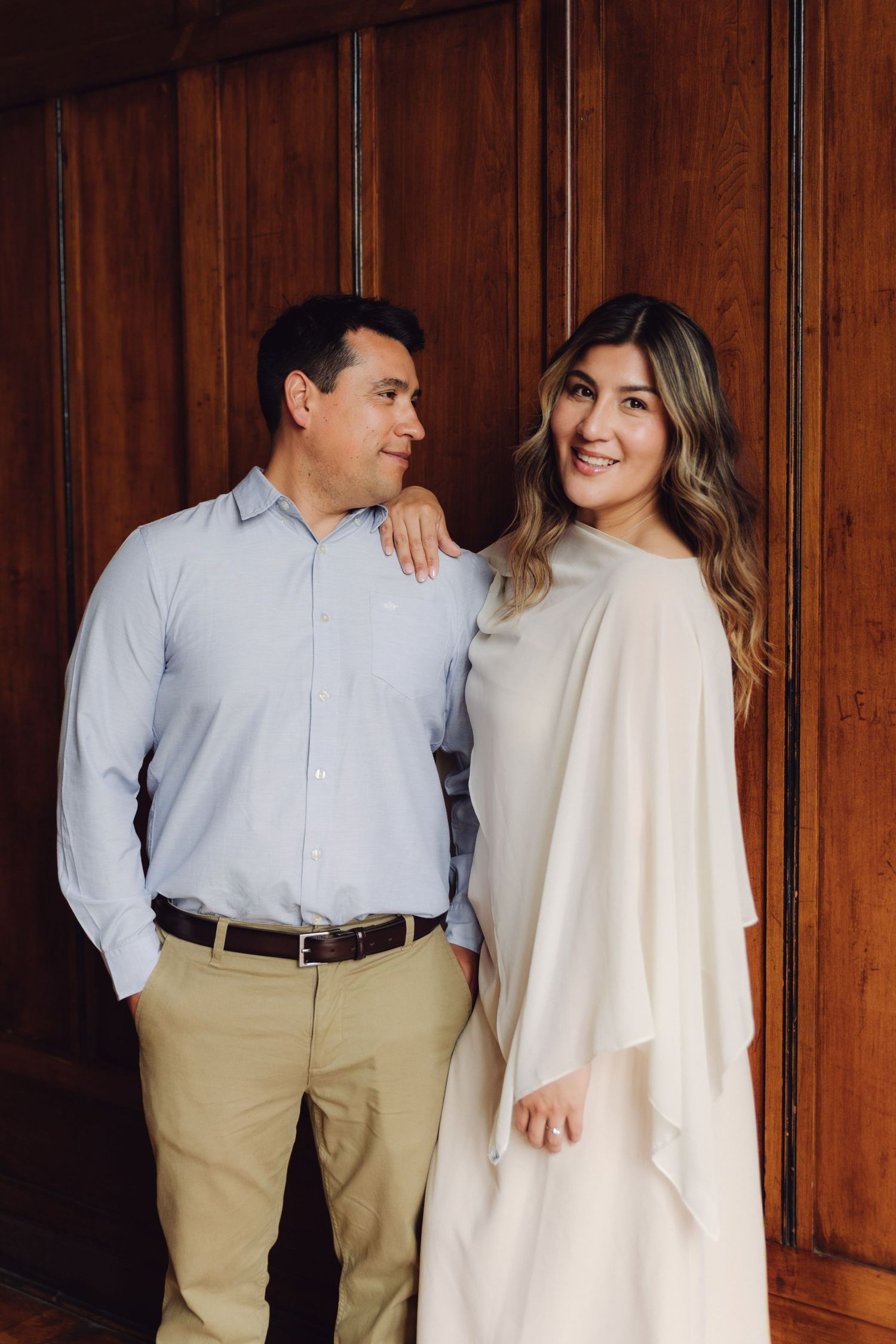Smiling couple posing together against warm wooden wall, woman in white dress with flowing cape.