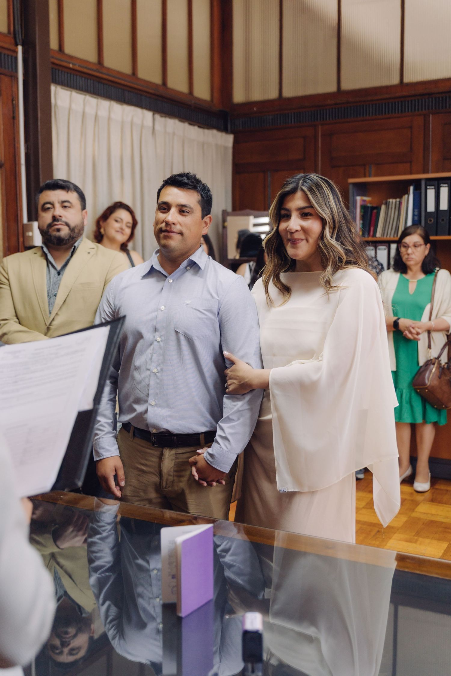 Smiling couple holding hands at a civil wedding ceremony with guests in a formal office setting.