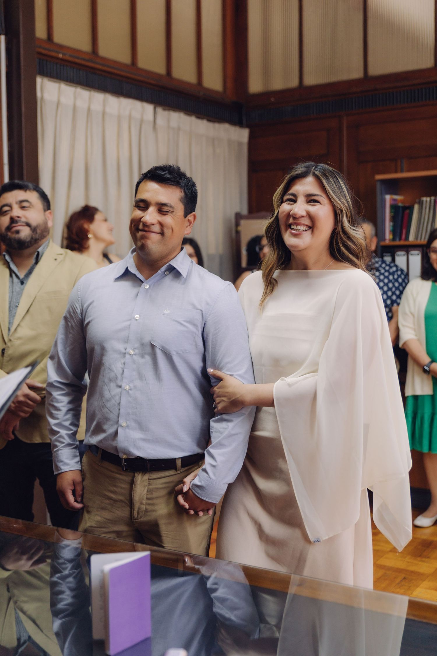 Happy couple holding hands and smiling during civil wedding ceremony with guests in background.