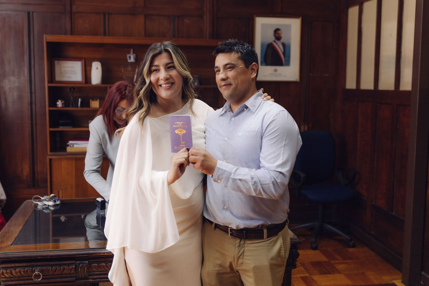 Smiling newlywed couple posing together holding their purple marriage certificate booklet indoors.
