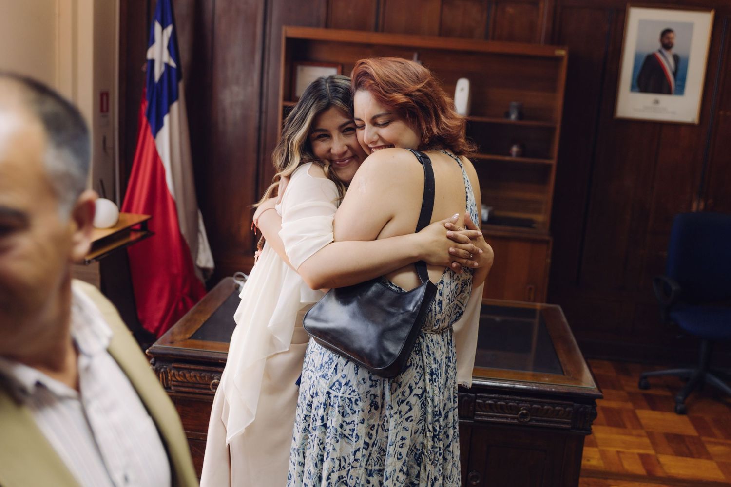 Bride embracing a female guest warmly with Chilean flag visible in the background after wedding.