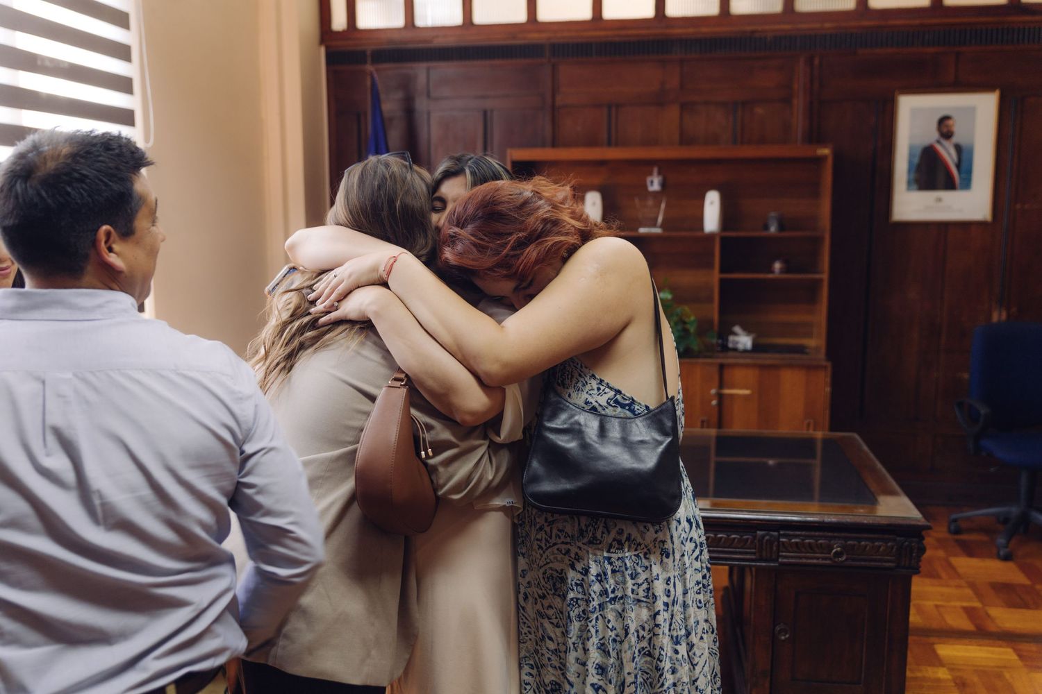 Three people share an emotional group hug in a formal office setting during a special celebration.