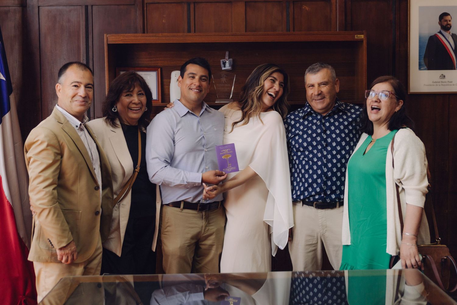 Six people pose together smiling in an official office, holding a document during a civil ceremony.