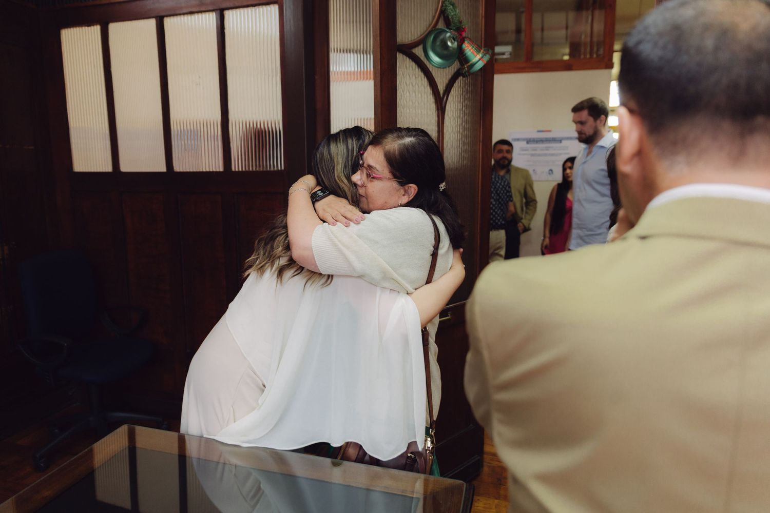 Two women hug affectionately inside a rustic wooden venue while guests look on nearby.