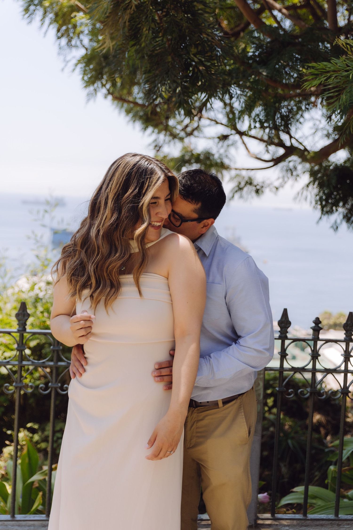 A man kisses a woman in a white dress on the cheek at a beautiful coastal viewpoint.