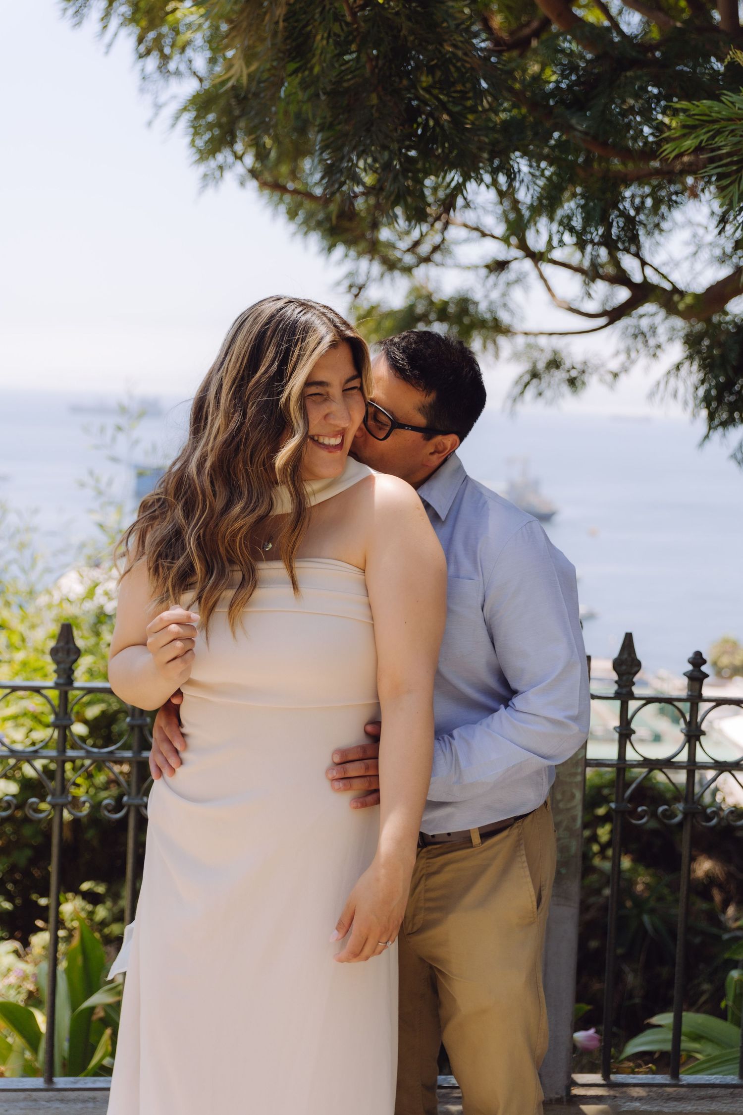A smiling couple embraces outdoors with a stunning ocean backdrop and lush greenery.