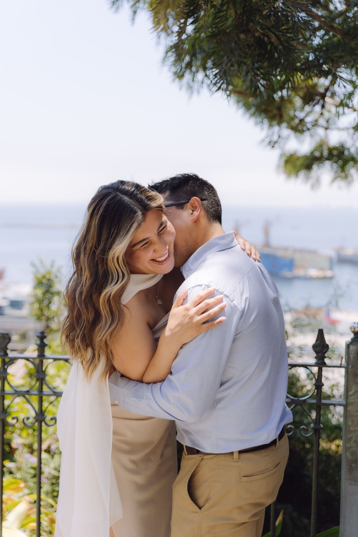 A laughing couple embraces warmly at a scenic outdoor terrace with ocean and port views.