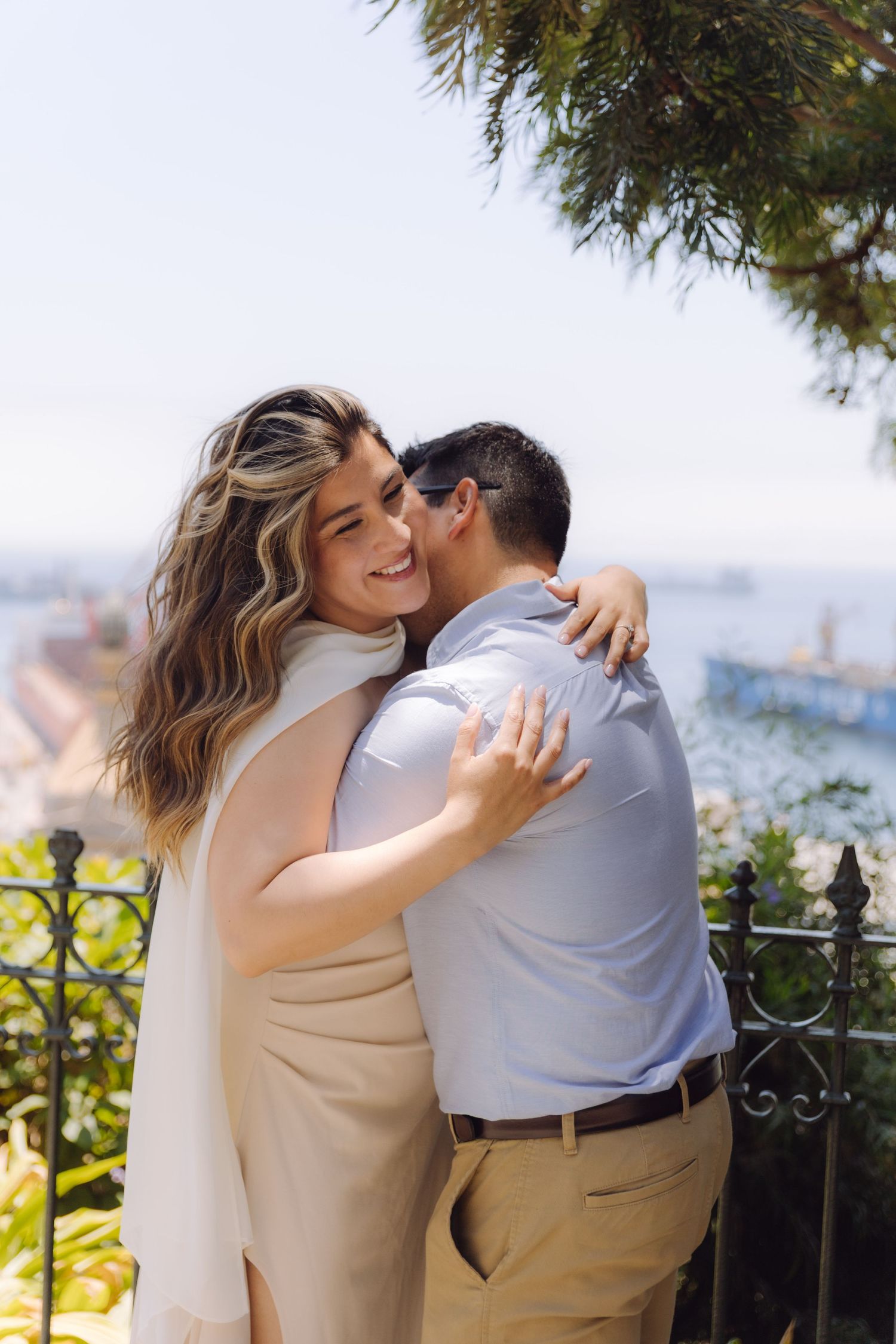Smiling woman with wavy hair hugs man outdoors with ocean view backdrop during engagement photo session.
