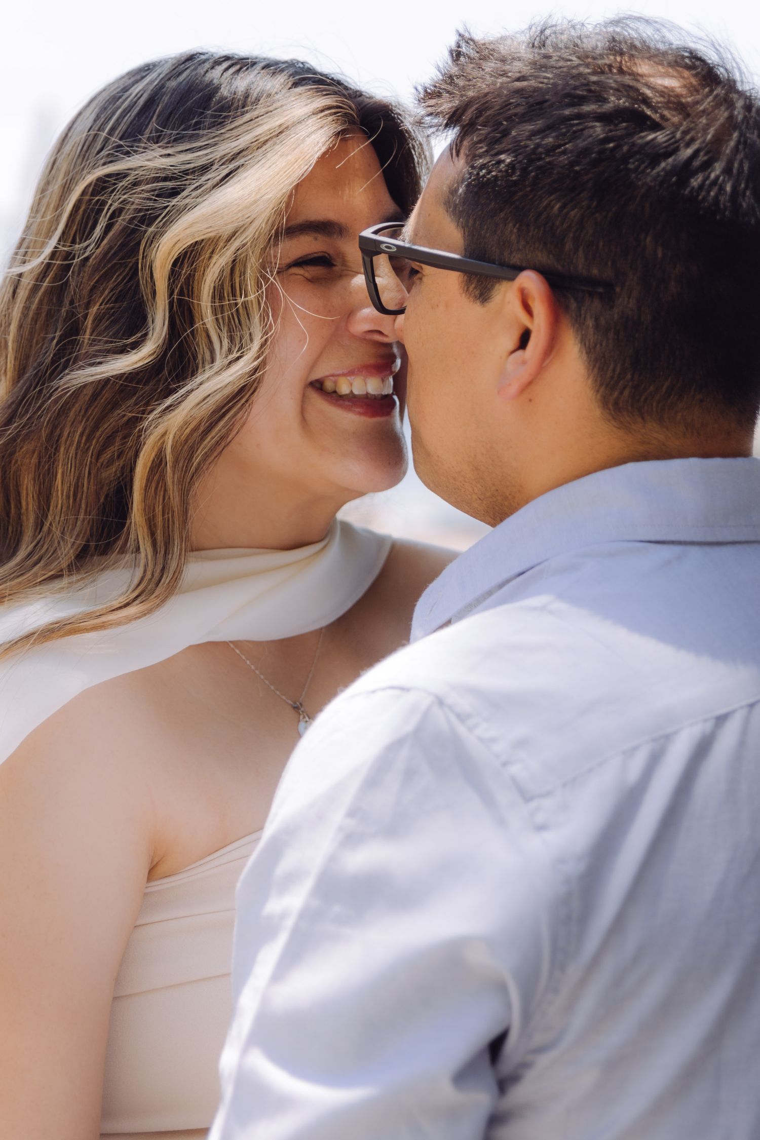 Close-up of laughing couple about to kiss, woman wearing glasses and white top in bright sunlight.