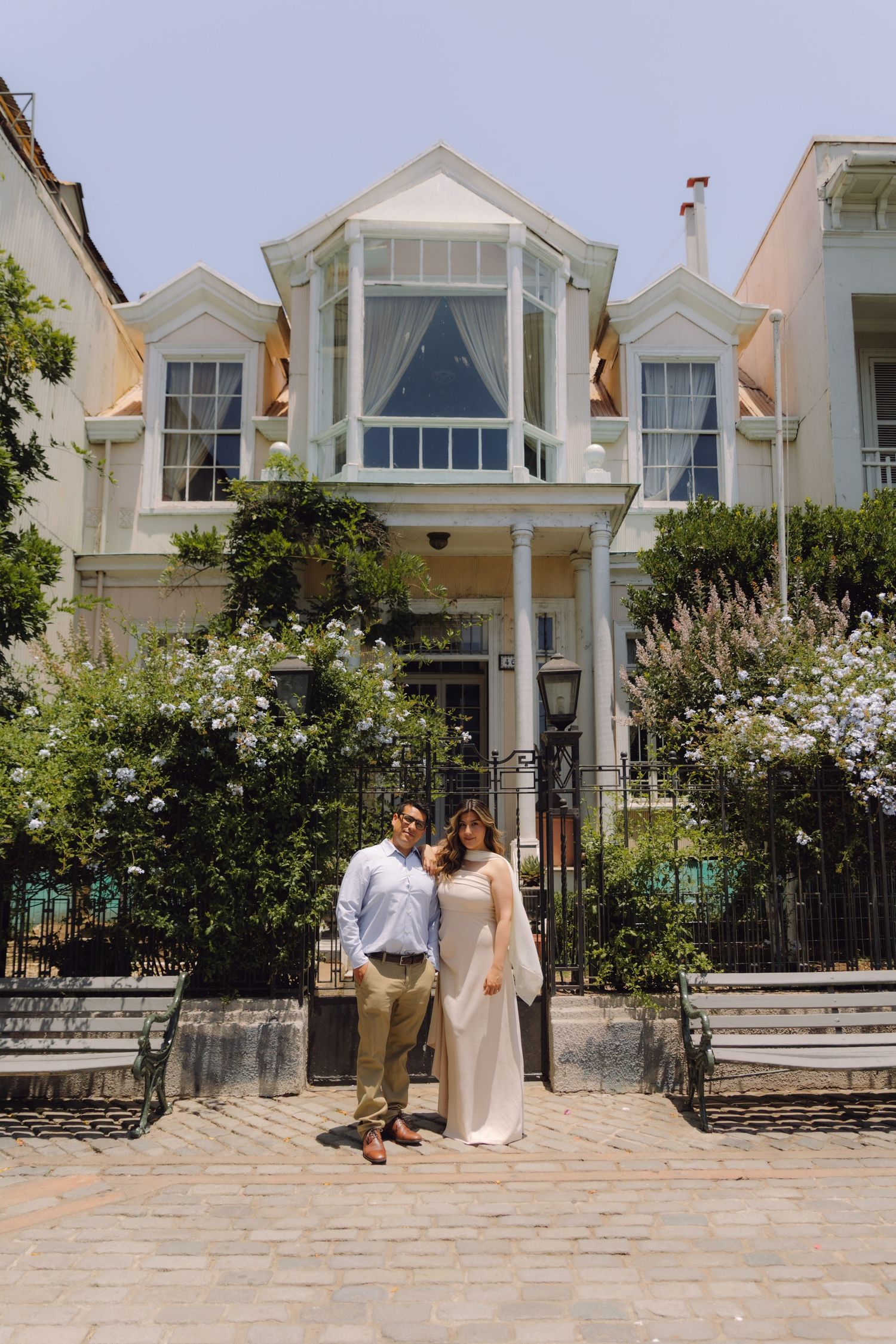 Couple in elegant attire standing together before charming white Victorian house with cobblestone path.