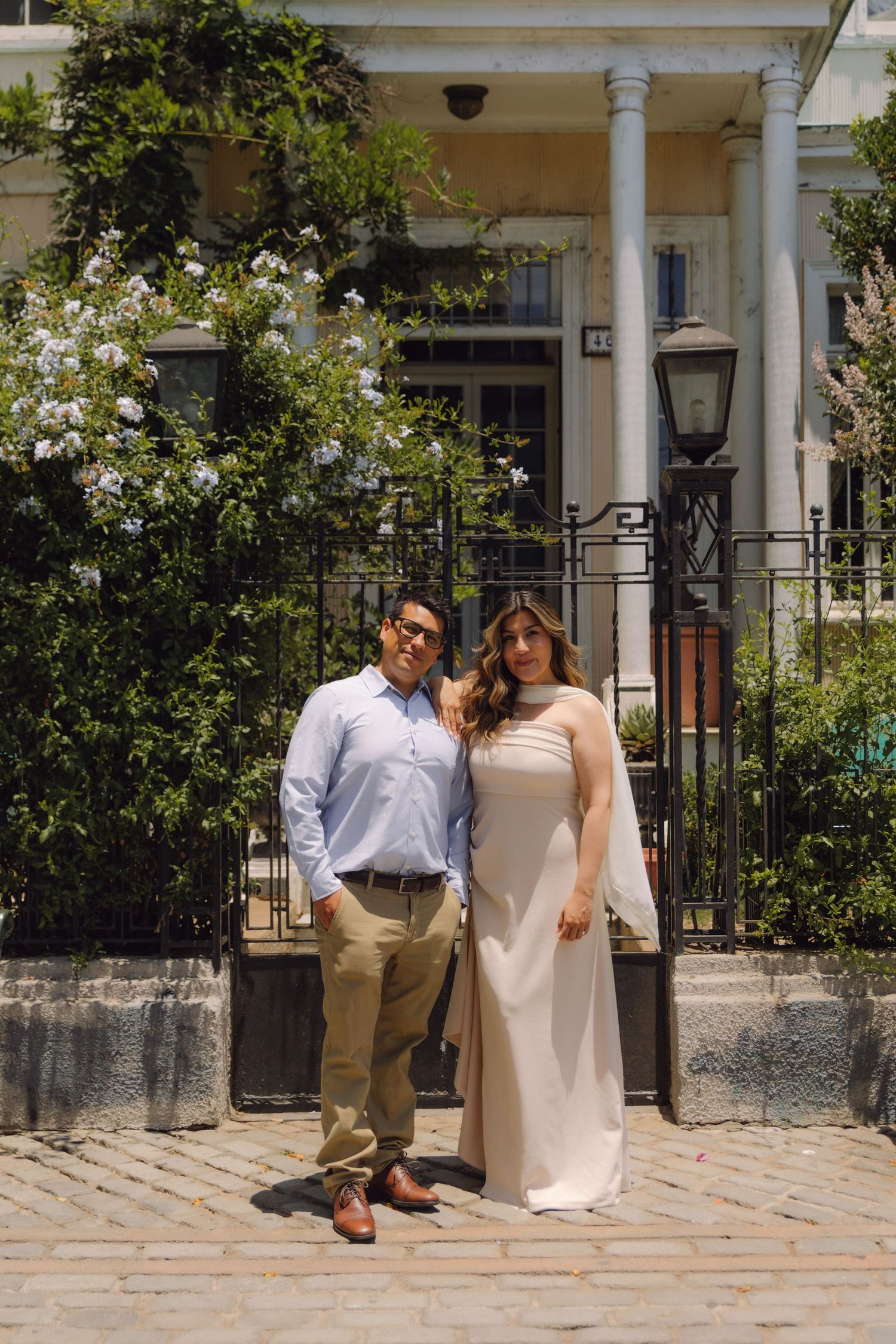 Stylish couple posing before ornate iron gate and white colonial building with blooming flowers.
