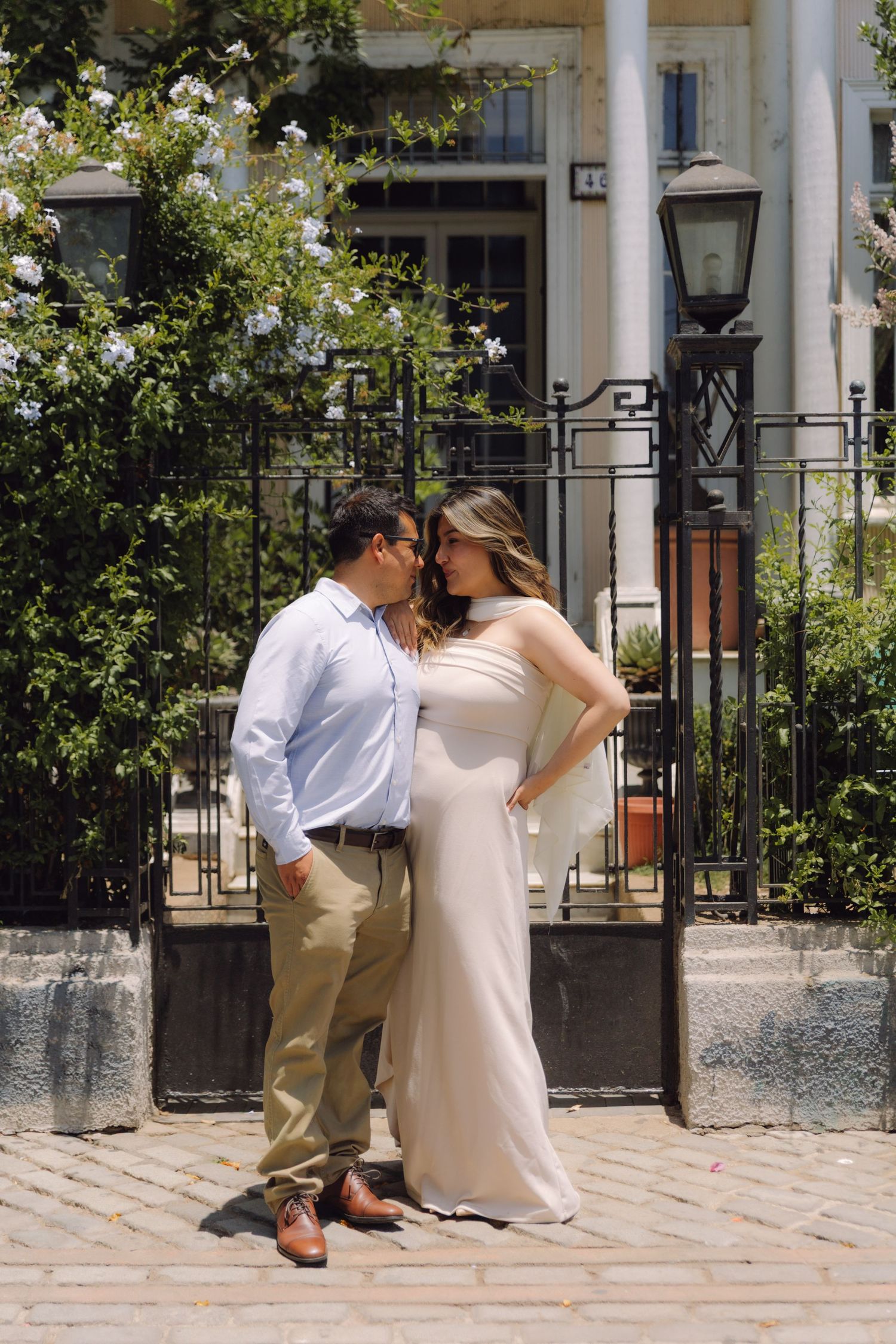 Romantic couple sharing tender moment by iron fence with white colonial architecture and lush greenery.